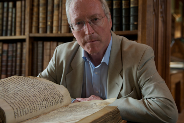 Diarmaid Macculloch, sat at a table in a library, looking up from an open manuscript.